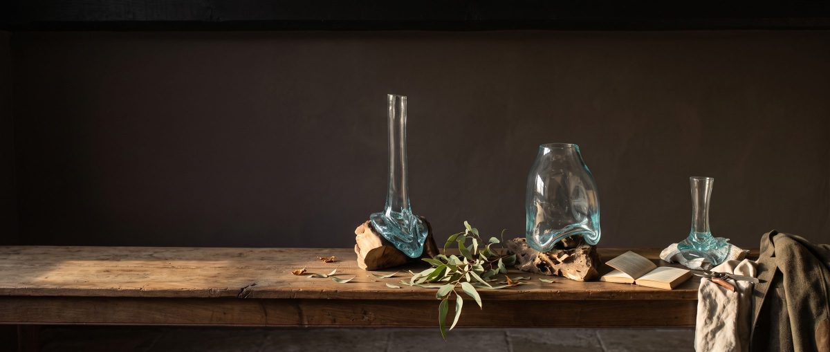 Trois vases en verre soufflé alignés sur un banc de bois, une branche d'olivier posée à côté