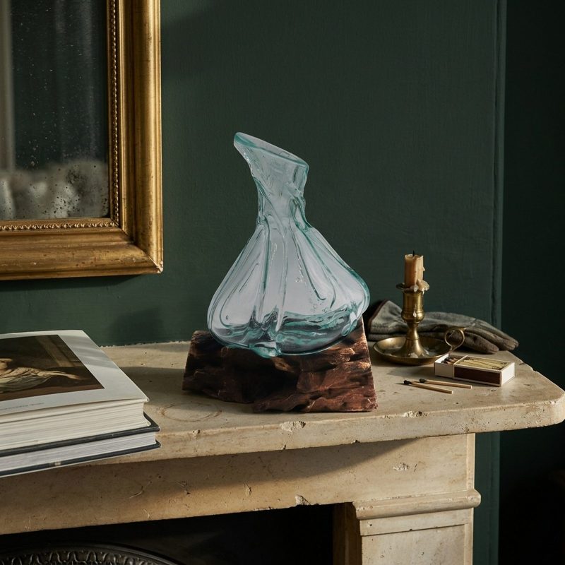 Petite carafe travaillée en verre soufflé posée sur un manteau de cheminée en pierre claire, bougeoir en laiton et boîte d'allumettes à côté, mur vert foncé