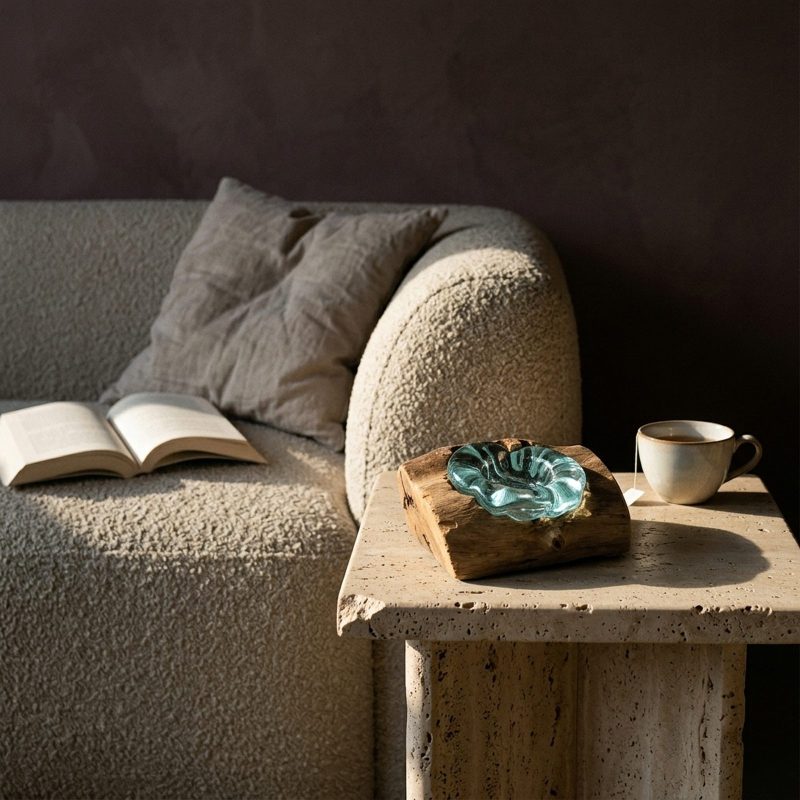 Photophore en verre soufflé posé sur un bout de canapé en travertin, tasse en grès à côté, canapé en bouclé et livre ouvert à l'arrière-plan